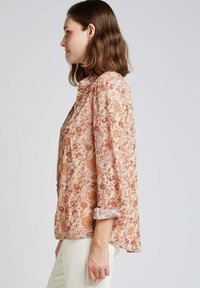 Blouse à manches longues avec un imprimé floral présentant des motifs orange et bordeaux sur un fond clair. Tissu doux et léger avec des poignets froncés.