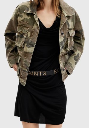 Kvinde iført en grøn camouflagejakke over en sort kjole med et nittebælte med teksten "SAINTS RO".