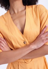 Žena se založenýma rukama, oblečená v lehkém oranžovém šatech s výstřihem do V, knoflíkem a detailem opasku, s přirozeným tónem pleti a kudrnatými vlasy.