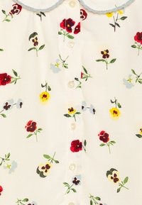 Hvit bluse med blomstertrykk i rød, gul og blå. Har en rund krage med lys blå kant og en knappeåpning foran.