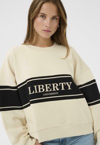 Sweat-shirt oversize crème avec une bande horizontale noire. "LIBERTY UNIVERSITY" imprimé en lettres crème en gras sur la bande. Matériau doux, encolure côtelée.