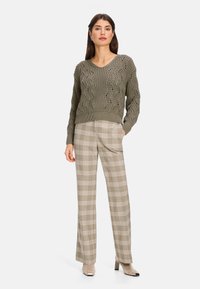 Olivgrüner gestrickter Pullover mit V-Ausschnitt und strukturiertem Muster, kombiniert mit beigen karierten Hosen. Das Model trägt hellfarbene Stiefeletten mit Absatz.