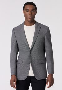 Grau karierter Blazer mit zwei vorderen Knöpfen, Reverskragen und aufgesetzten Taschen. Getragen über einem hellen Pullover. Glatter, strukturierter Stoff.