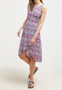 Kvinna i en ärmlös knälång klänning med rosa och lila cirkulära mönster samt asymmetrisk fåll, kombinerad med grå sandaler.