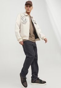 Jauns vīrietis beigu beisbola jaciņā, brūnā T-kreklā, melnās biksēs un melno sporta apavu pozē pret baltu fona.