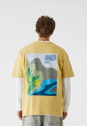 Tricou galben din bumbac cu un grafic colorat al Coastei Amalfi pe spate, prezentând munți, apă și accente verzi.