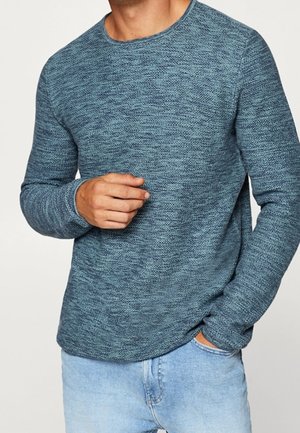 Man met een blauw gestructureerd trui met lange mouwen en lichtblauwe spijkerbroek, met één hand in de zak en de andere die de borst raakt.