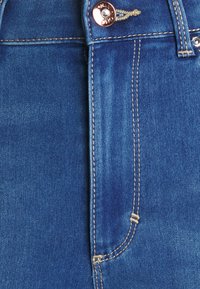 Jeans de mezclilla azul con una cintura alta, botón de cobre y costuras doradas en contraste a lo largo de las costuras y los bordes de los bolsillos.
