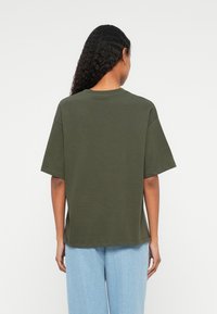 Ελαιοπράσινο βαμβακερό t-shirt με χαλαρή εφαρμογή, κοντά μανίκια και στρογγυλή λαιμόκοψη. Το ύφασμα έχει λεία υφή και ελάχιστες ραφές.