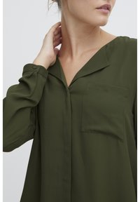 Blusa verde oliva con scollo a V, maniche lunghe, polsini buttonati e taschino sul petto. Realizzata in tessuto liscio con una vestibilità comoda.