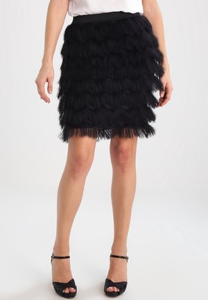 Mini-jupe noire à franges superposées portée avec un haut blanc et des talons noirs à bride à la cheville sur fond uni.