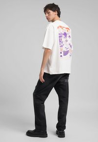 T-shirt blanche surdimensionnée avec un graphique violet et orange au dos, associée à un pantalon en denim foncé et des chaussures noires.