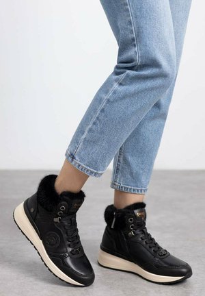 Persoon draagt lichtblauwe spijkerbroek en zwarte hoge sneakers met bontvoering en witte zolen op een grijze vloer.