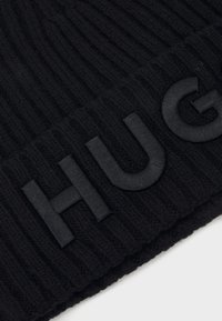 Beanie lavorato a maglia nero con texture a coste e risvolto. Presenta un logo "HUG" in pelle nera ben visibile sul davanti.
