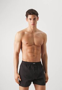 Zwarte katoenen boxershorts met een elastische tailleband en een knoopsluiting. Model staat zonder shirt en toont een eenvoudig ontwerp.