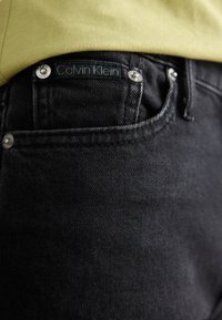 Juodos džinsai turi Calvin Klein etiketę ant galinio kišenių, sidabrinius aksesuarus ir tekstūruotą audinio apdailą.