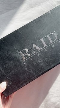 Boîte à chaussures rectangulaire noire avec une finition mate, comportant un logo argenté en relief "RAID LONDON" sur le dessus. Bords nets, peu d'usure.