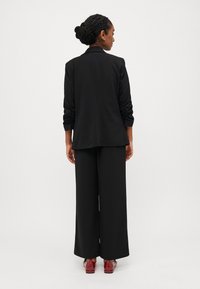 Μαύρο ταγέρ με ρυμουλκούμενα μανίκια, συνδυασμένο με χαλαρές μαύρες παντελόνες. Το μοντέλο φοράει κόκκινα παπούτσια, απεικονίζεται από πίσω σε απλή φόντο.