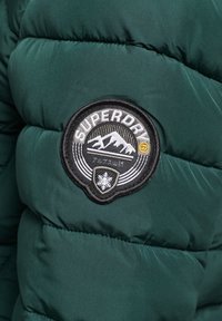Tela de chaqueta acolchada verde con un parche circular en blanco y negro que dice "Superdry." El parche presenta diseños de montañas y copos de nieve.