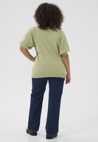 Groen kort mouwshirt met een losse pasvorm en geribbelde zoom, gecombineerd met donkerblauwe wijde jeans en zwarte schoenen. Krullend haar zichtbaar.