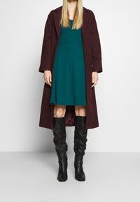Manteau bordeaux mi-genoux porté sur une robe bleu canard, assorti à de grandes bottes noires texturées. Tissu lisse et lignes simples et épurées.