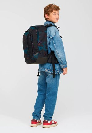 Ragazzo con giacca di jeans e jeans che indossa uno zaino grande nero e blu, in piedi di lato e sorridente su sfondo semplice.