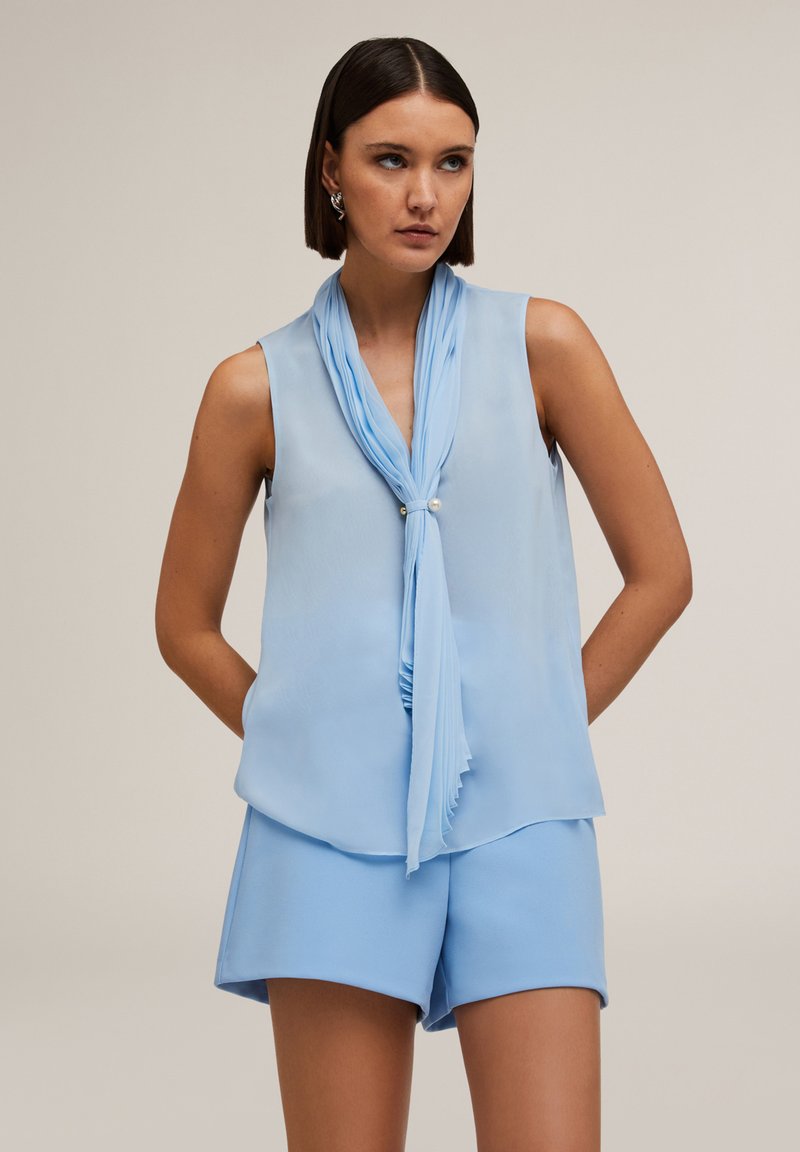 Jasnoniebieska bluzka bez rękawów z detalem w postaci plisowanego szalika i perłowym akcentem. Miękka, zwiewna tkanina i luźny krój, połączona z dopasowanymi szortami.