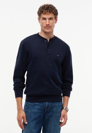 Man draagt een marineblauw henley-trui en blauwe spijkerbroek, staand met een neutrale expressie tegen een witte achtergrond.