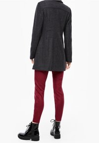 Donna che indossa un cappotto grigio scuro lungo fino al ginocchio, pantaloni rossi attillati e stivaletti neri lucidi con lacci, in piedi con la schiena rivolta in avanti.