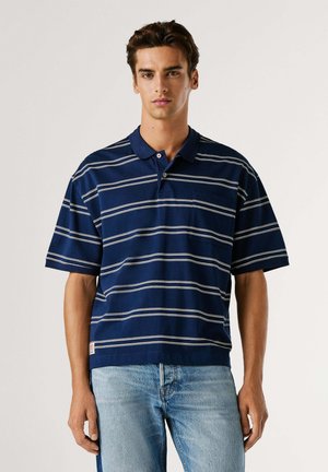 Marineblauw poloshirt met witte strepen, korte mouwen en een kraag. Voorzien van een borstzak en geribbelde zoom. Gecombineerd met lichtblauwe spijkerbroek.