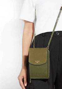 Green crossbody bag with textured and smooth surfaces, zip closure, logo on front, and a woven strap. Compact rectangular shape.