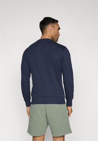 Felpa girocollo blu navy con maniche lunghe, polsini e fondo a coste. Indossata con pantaloni corti verde chiaro, design minimalista, texture del tessuto morbida.