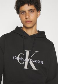 Melnā kapucīna jaka, kas izgatavota no mīkstas fabric, ar lielu "CK" logotipu un "Calvin Klein Jeans" tekstu baltā un pelēkā krāsā priekšpusē.