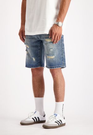 Denim shorts, lichtblauw met versleten details en een rafelige zoom. Gecombineerd met witte sokken en zwartgestreepte sneakers. Witte t-shirt.
