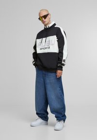 Sort og hvid hoodie med "FUBU SPORTS" tryk, der har høj krave og brede ærmer; parret med løse blå jeans og hvide sneakers.