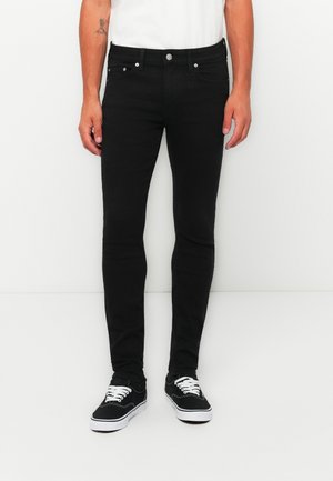 Homme portant un jean noir slim, un t-shirt blanc et des baskets noires avec des lacets blancs, debout devant un fond blanc uni.
