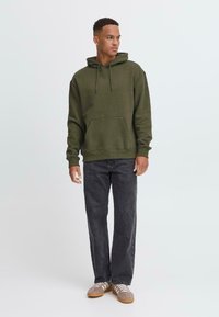 Olijfgroen hoodie met een voorzak, trekkoordkap, en geribbelde manchetten, gecombineerd met donkergrijze recht-pijp jeans en lichtbruine sneakers.