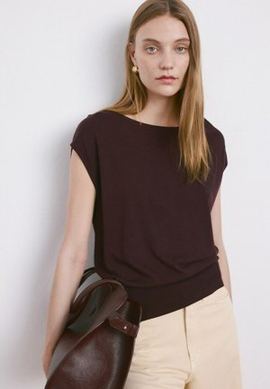 Jeune femme aux longs cheveux portant un haut sombre sans manches et un pantalon clair, tenant un grand sac à main en cuir marron foncé à son côté.