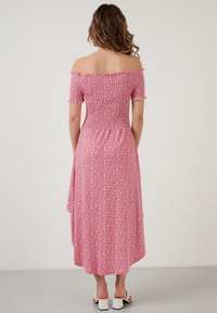 Vaaleanpunainen kukallinen olkaimeton mekko, jossa on röyhelöhihoja, kootut rintatukikappaleet ja virtaava helma. Mukana on sivuaukko ja pehmeä, teksturoitu materiaali.