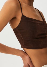 Vrouw in een aansluitend bruin cropped tanktop met dunne bandjes, hand op haar taille, te zien van schouders tot heupen.