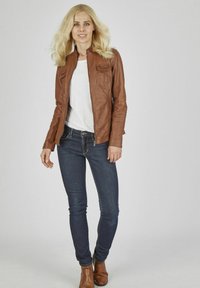 Veste en cuir marron zippée avec deux poches avant, portée sur une chemise blanche et un jean en denim foncé, complétée par des bottines marron.