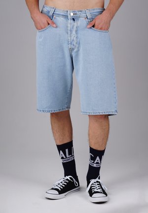 Hellblaue Jeansshorts mit geradem Schnitt, zwei Fronttaschen und einem gestickten Saum. Kombiniert mit schwarzen Turnschuhen und marineblauen Socken mit weißem Schriftzug.