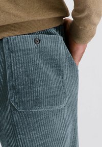 Man in een blauwe corduroy broek met een knoopzak aan de achterkant, hand in de zak, en een bruine gebreide trui.