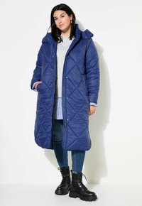Abrigo puffer largo acolchado azul con capucha, que presenta botones a presión, bolsillos laterales y costuras en patrón de diamante, llevado sobre una camiseta gris y jeans.