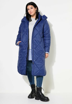 Blue quilted long puffer coat with a hood, featuring snap buttons, side pockets, and diamond pattern stitching, worn over a gray top and jeans.