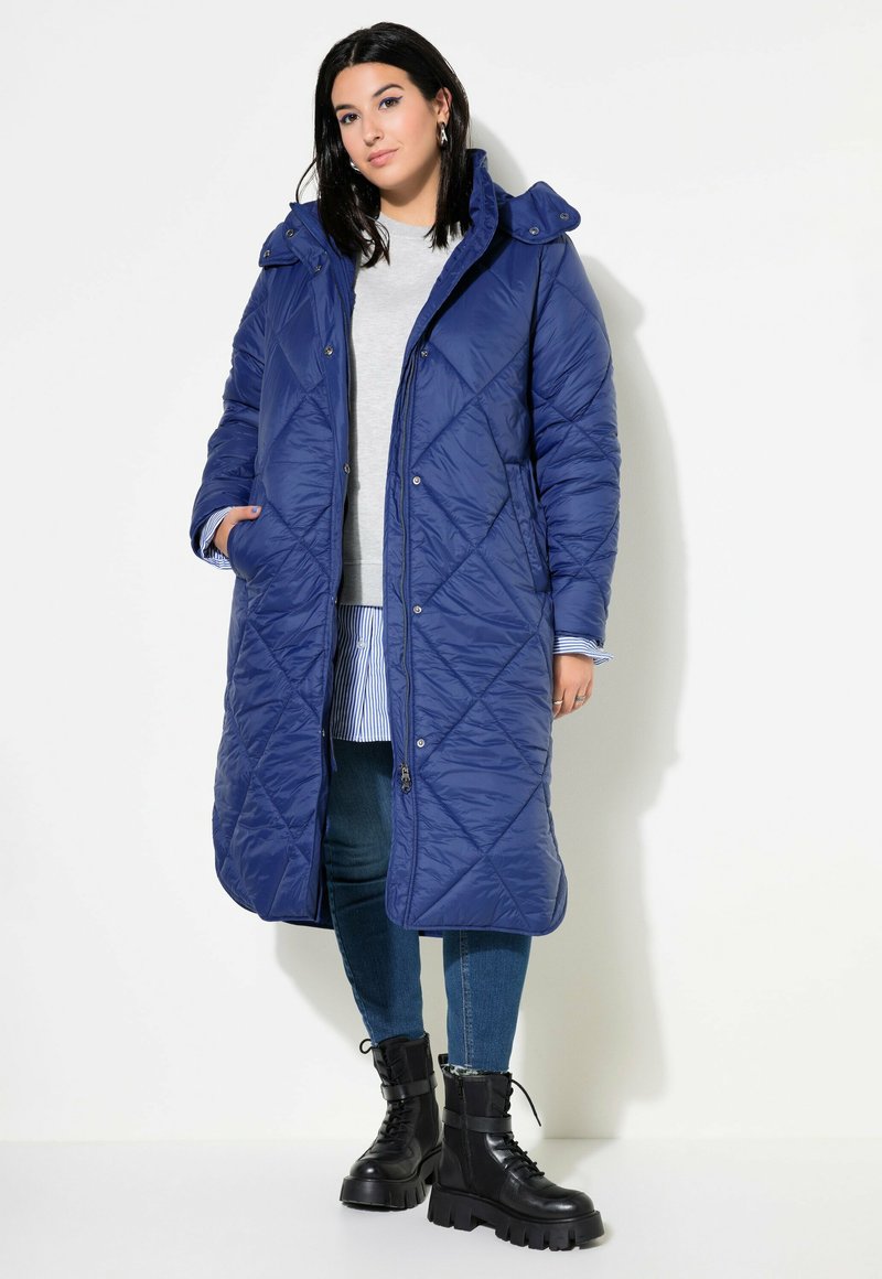 Abrigo puffer largo acolchado azul con capucha, que presenta botones a presión, bolsillos laterales y costuras en patrón de diamante, llevado sobre una camiseta gris y jeans.