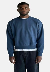 Felpa blu scuro con colletto rotondo, maniche lunghe e taglio cropped, indossata sopra una maglietta bianca. Il tessuto sembra liscio.