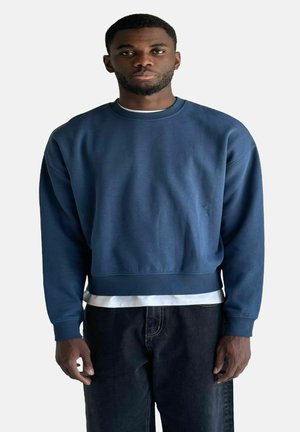 Dunkelblaues Sweatshirt mit rundem Ausschnitt, langen Ärmeln und kürzerer Länge, über einem weißen T-Shirt geschichtet. Der Stoff wirkt glatt.