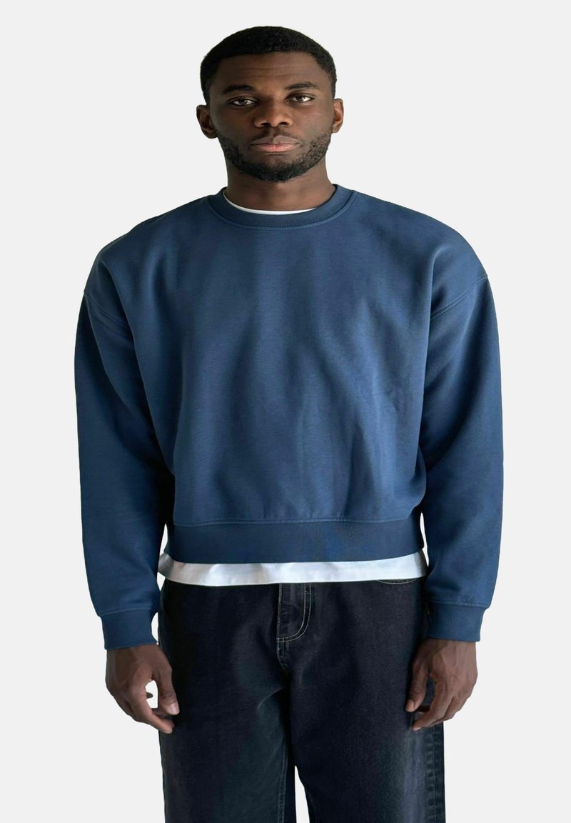 Felpa blu scuro con colletto rotondo, maniche lunghe e taglio cropped, indossata sopra una maglietta bianca. Il tessuto sembra liscio.