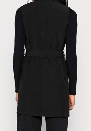 Osoba nosząca długą czarną kamizelkę bez rękawów na czarnym topie z długim rękawem, pokazana od tyłu z paskiem w talii.